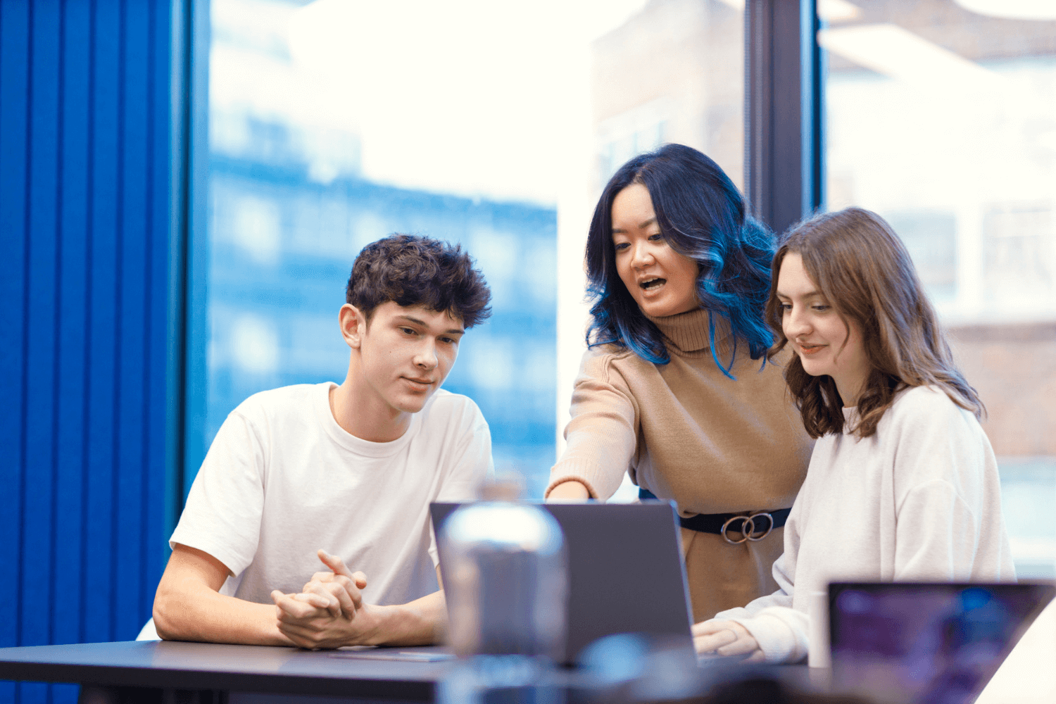 A group of three people gathered around a laptop. A woman in a tan coloured jumper with long brown hair and blue highlights, is explaining something to two younger colleagues. One colleague is male and one is female; both are wearing white tops. 