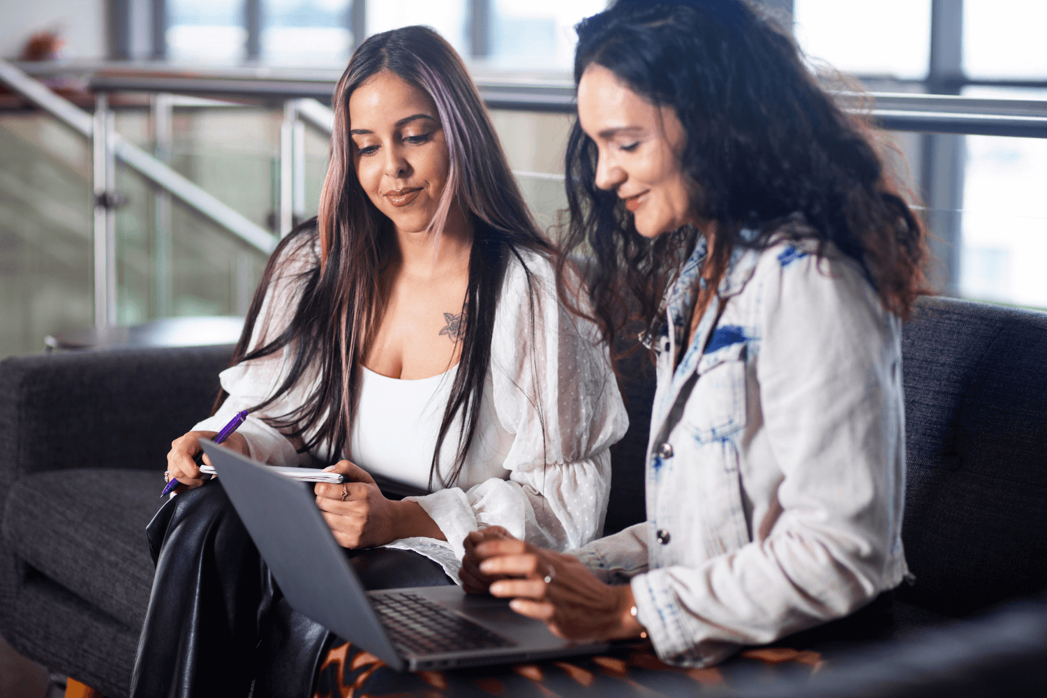Two women are working together. They are sat on informal seating, looking at a laptop. One woman has dark curly brown hair and wears a denim jacket. The other has long straight brown hair, with pink highlights.  She has a long white top with baggy sleeves, and a tank top underneath. 
