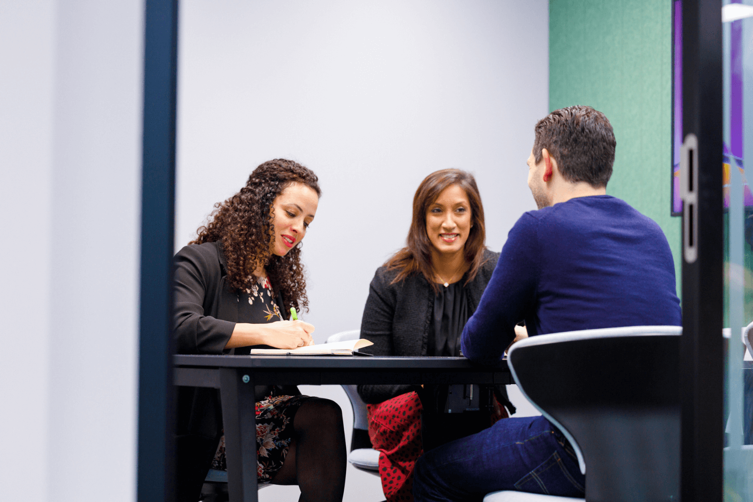 Three colleagues having a meeting.The meeting room is green, with a table in the middle. There are two women facing forward, and one man with his back to the camera.