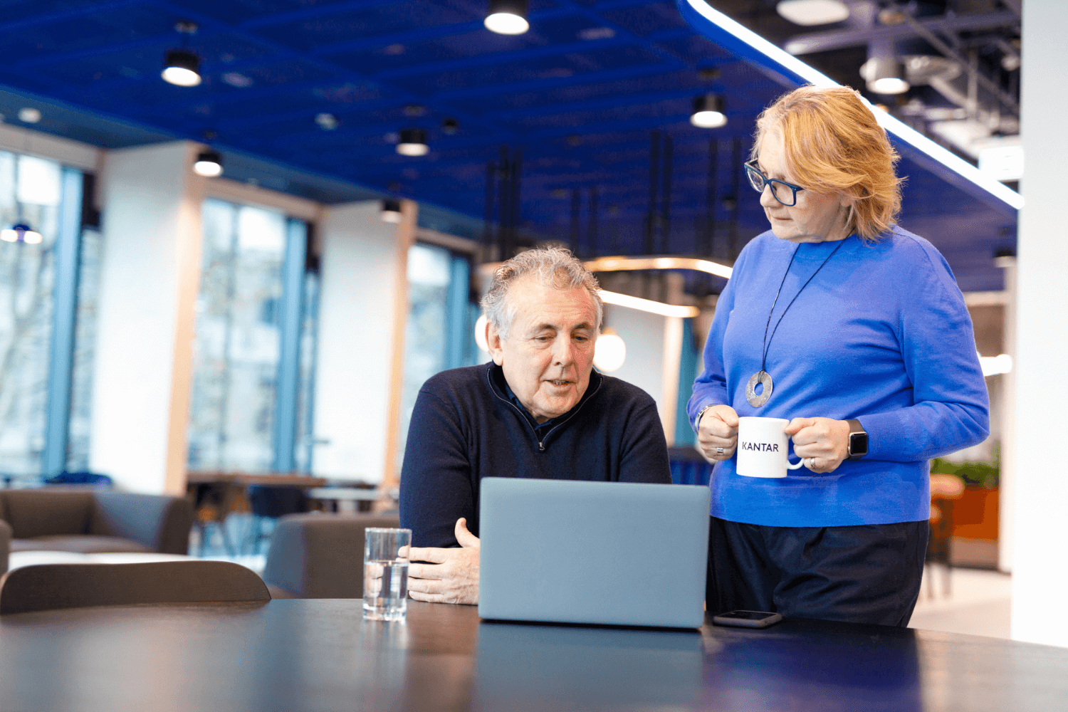 A woman in a blue jumper is stood up and holding a coffee. At the table, is a man in black jumper with his arms crossed, looking at a laptop.