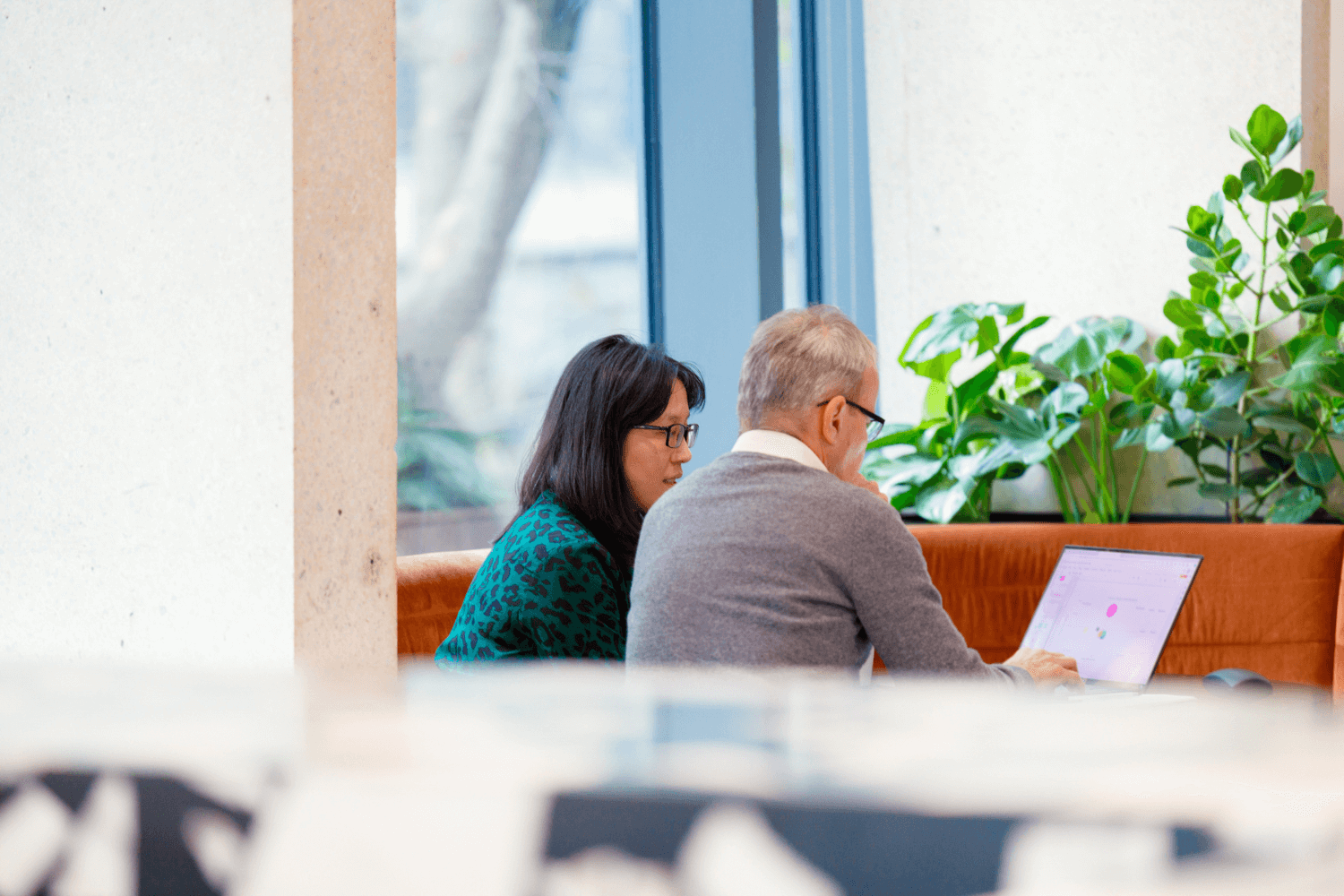 Two colleagues working together from a laptop. She is has shoulder length dark hair, and wears glasses and a green dress. He is wearing a mid-grey jumper and white shirt. 