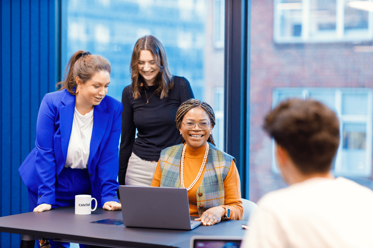 Four colleagues having a meeting together. There are three women and one man. One woman is working from a laptop and smiling at the camera, the others are standing around her, looking at the screen.