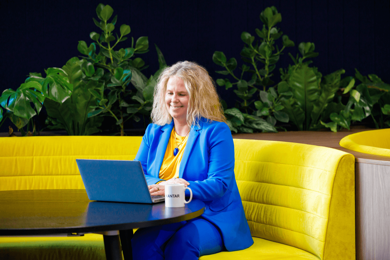 A woman with long curly blonde hair sat working at a laptop on a yellow sofa.