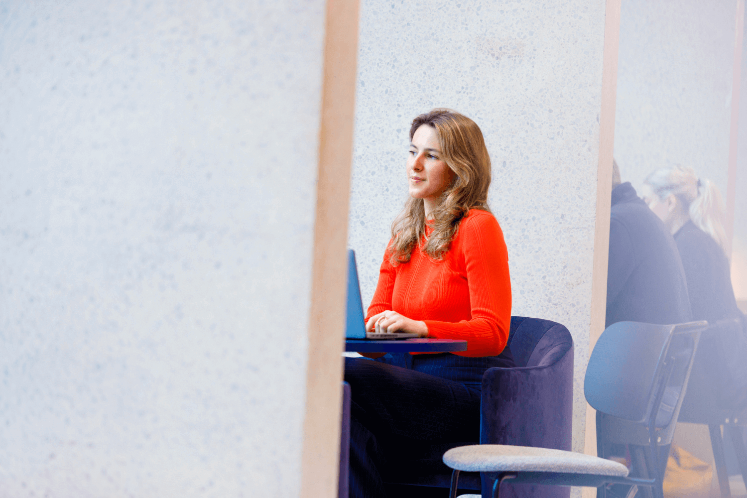 A young woman works at a table in a hidden alcove. She has light brown hair and is wearing an orange top.