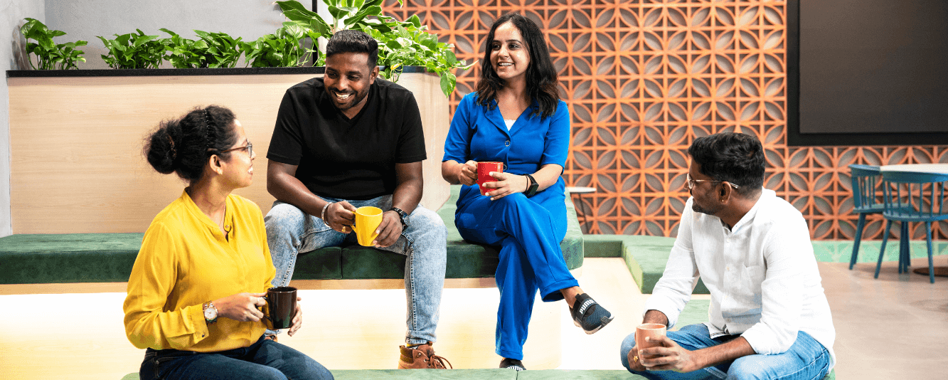A group of four colleagues are relaxing in the breakout area. They are sat on green velvet steps on different levels. At the highest level is one man and one woman. He is wearing a black T-Shirt, light blue jeans and is holding a yellow mug. His female colleague is wearing a blue jumpsuit and has a red mug. On the lower level there is a female colleague wearing a yellow blouse and holding a black mug. Finally, there is a male colleague in a white shirt and jeans; he is looking up at those sat above him. 