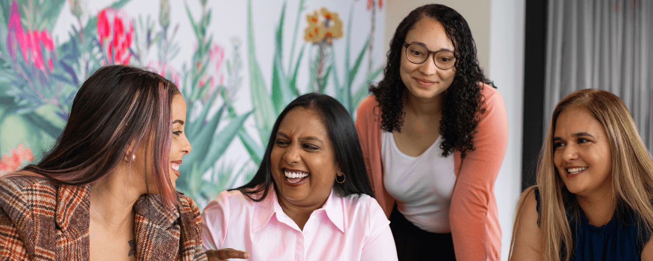 A group of four women are laughing together. From the left: the first woman has long brown hair with pink streaks in the front. She is wearing a brown and orange tweed-esque jacket. The next woman is wearing a pink shirt and has long brown hair, with gold hoop earrings. She is looking at the first woman. The third woman has on a plain white top and pink cardigan. She has long brown curly hair and wears glasses. The final woman has long blonde hair and is wearing a short-sleeved black top.