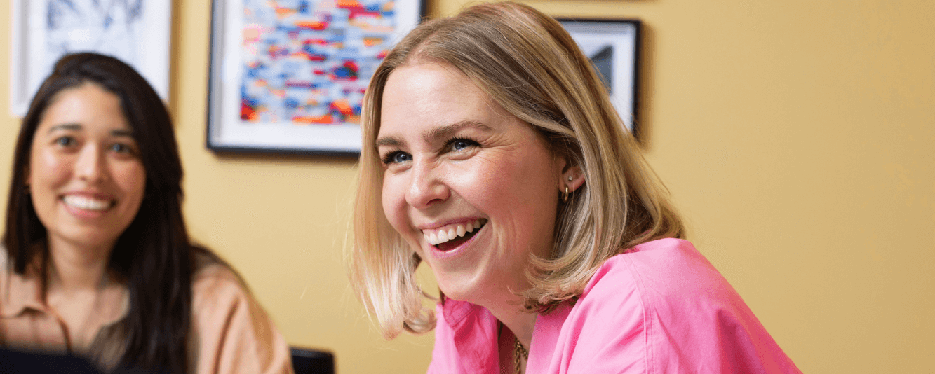 A woman with short blonde hair and wearing a pink shirt is laughing at the camera. She is sat in a room that is painted bright yellow. Behind her is another female colleague, slightly out of focus. She has long brown hair and is wearing a brown top.