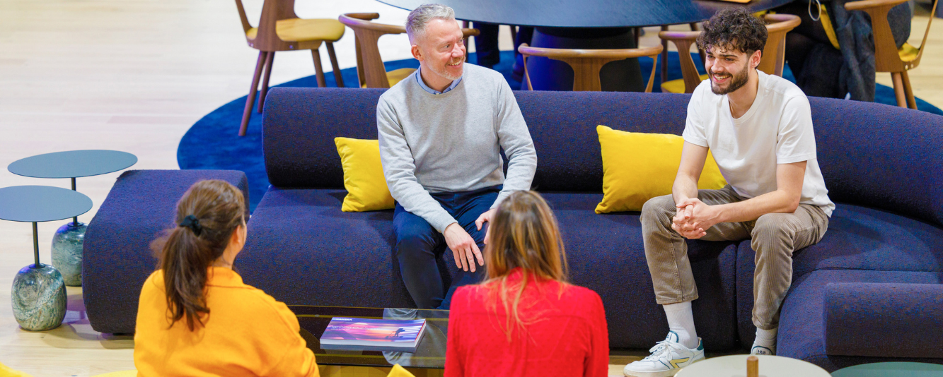 A group of four people sat together in a circle. Two males are sat on a dark blue sofa with yellow cushions, facing the camera. One is older in a light grey jumper and dark blue jeans. The younger has dark brown hair and is  a white T-shirt and khakhi trousers. Oppostite them are two women, both with brown hair. One is in a red shirt, the other in an orange jumper. They are facing away from the camera and you cannot see their faces.  at a laptop. 