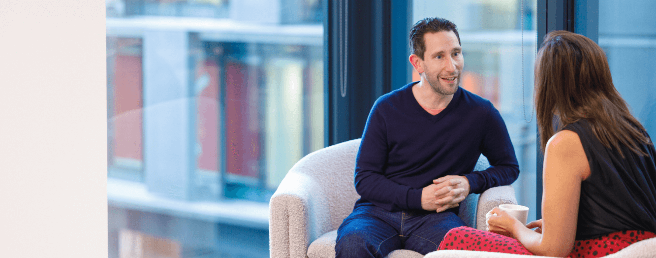 A man having a conversation with a female colleague, whilst sat on whit comfy chairs.