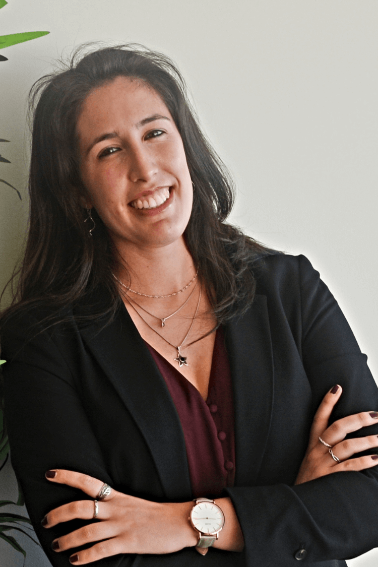 A woman leaning next to a plant. She has long brown hair and is wearing a black jacket and red dress. 