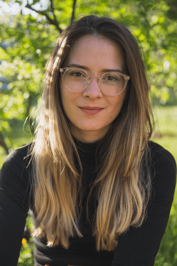A woman wearing a black polo, with long light brown hair and glasses. She is outdoors.
