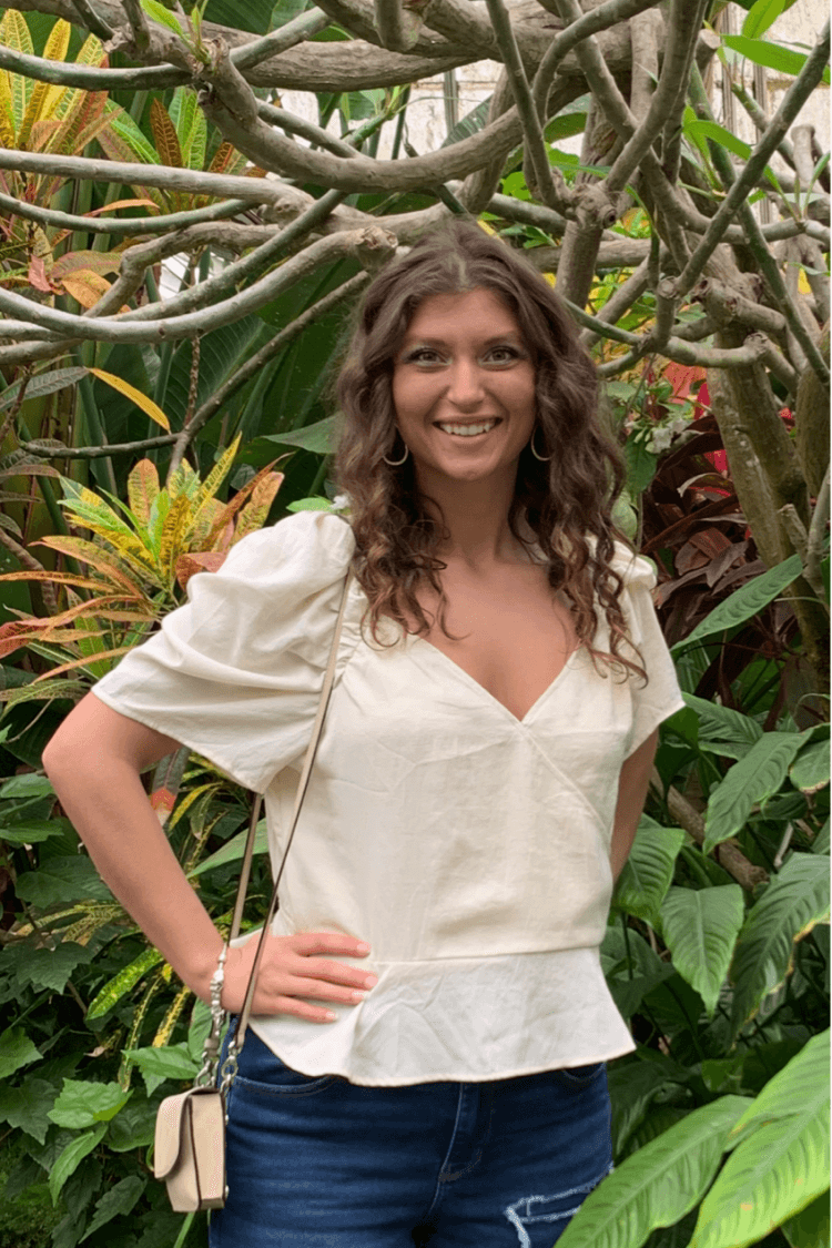 A woman in a white short sleeved top and jeans. She is surrounded by plants.