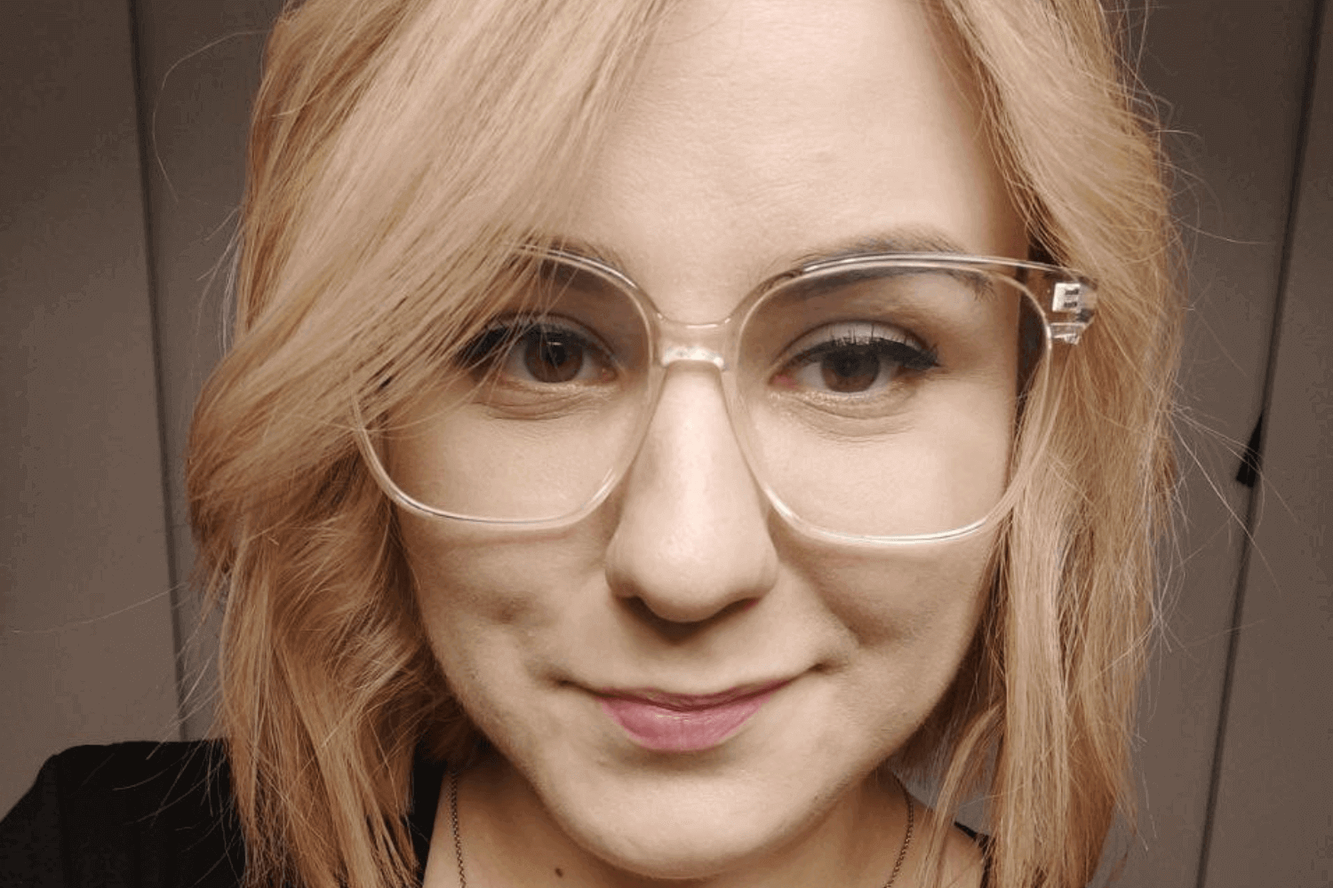 A woman with a black top and glasses with transparent frames. She has shoulder length blonder hair. 