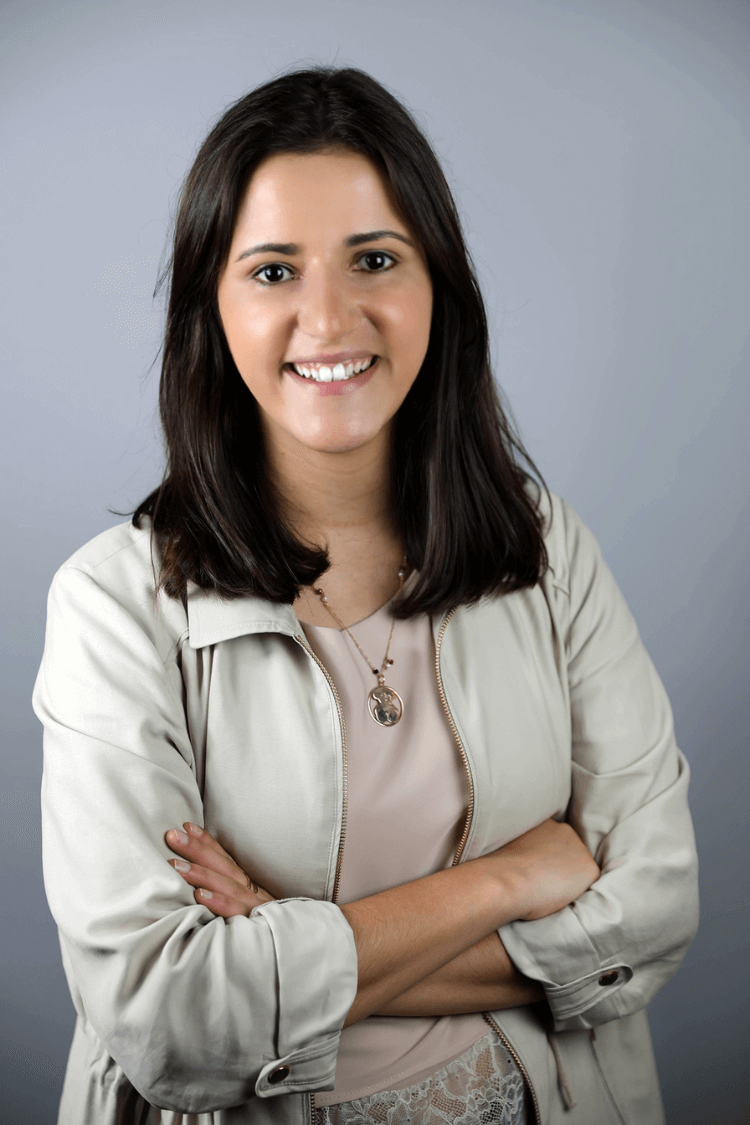 A woman smiling at the camera with her arms crossed. She is wearing a cream jacket and has shoulder length brown hair. 