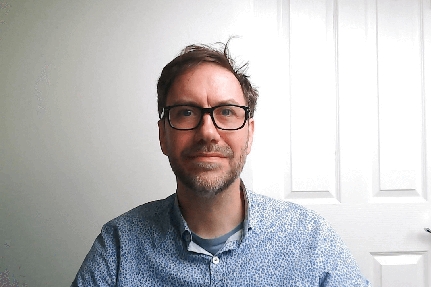 A man with black rimmed glasses looking at the camera, with a blue shirt on. He has short brown hair. 