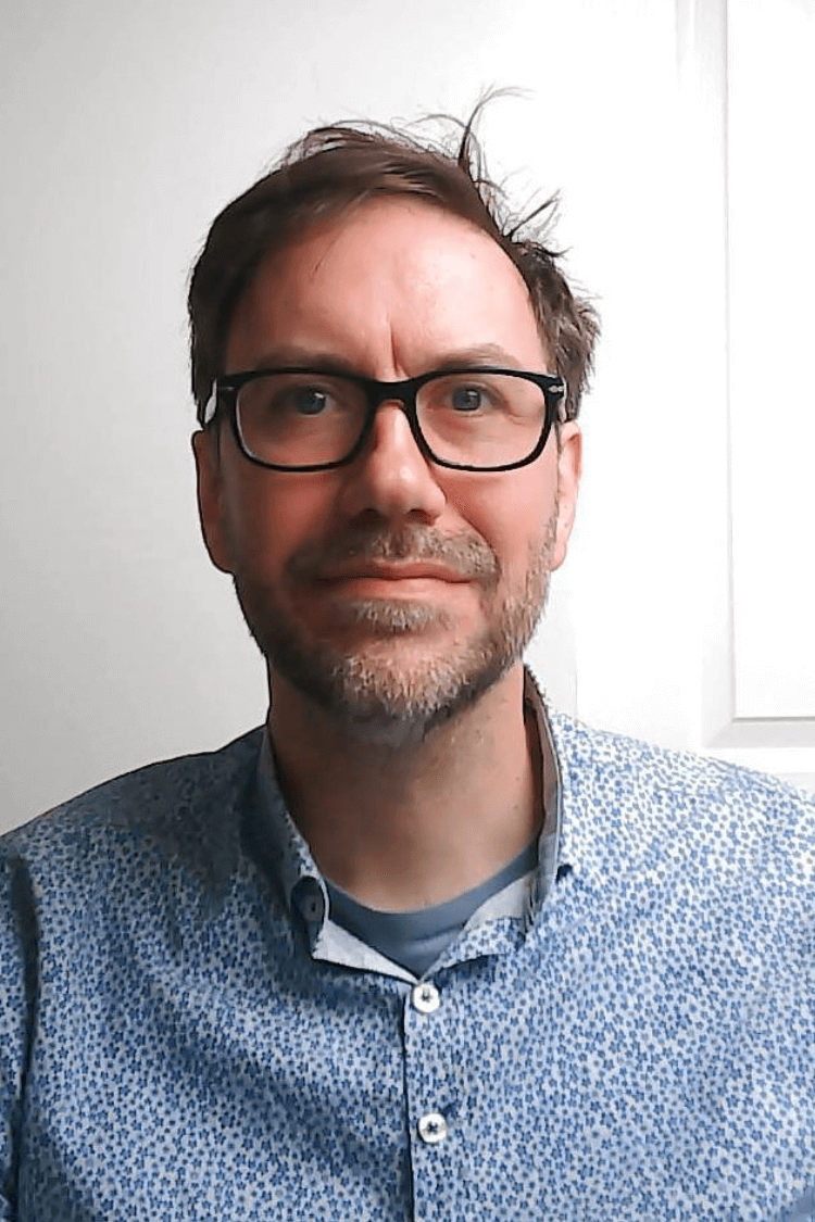 A man with black rimmed glasses looking at the camera, with a blue shirt on. He has short brown hair. 