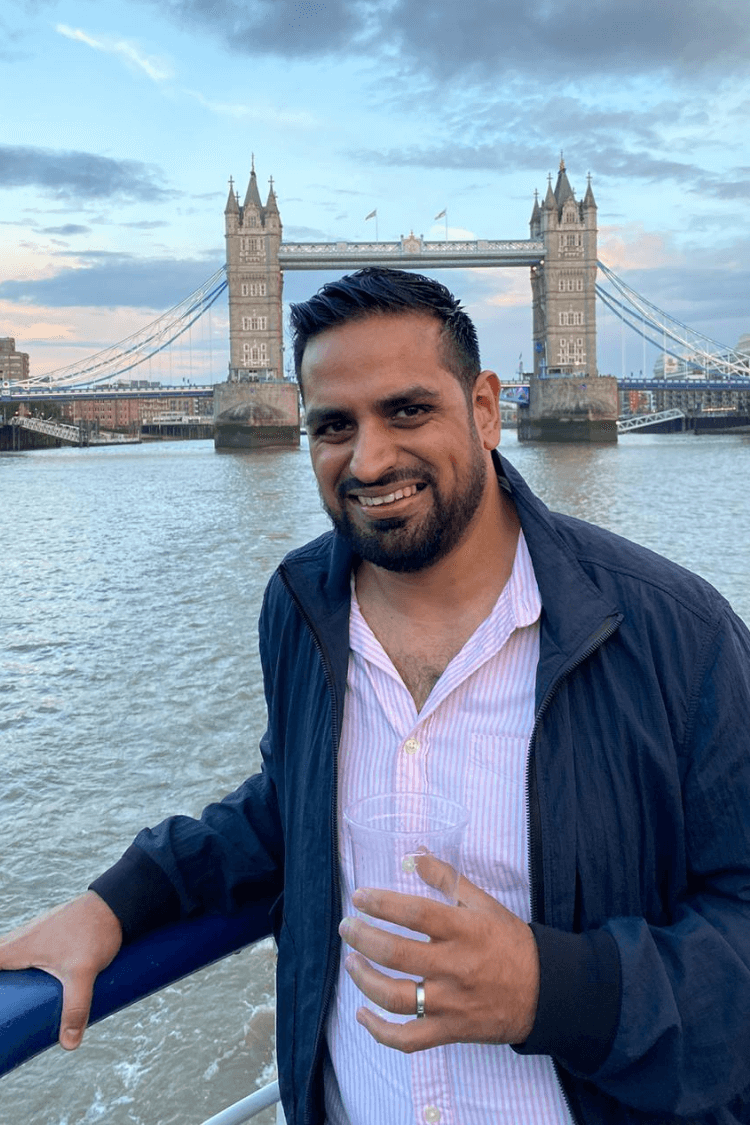 A man with a blue jacket and light shirt. Tower Bridge is behind him. 